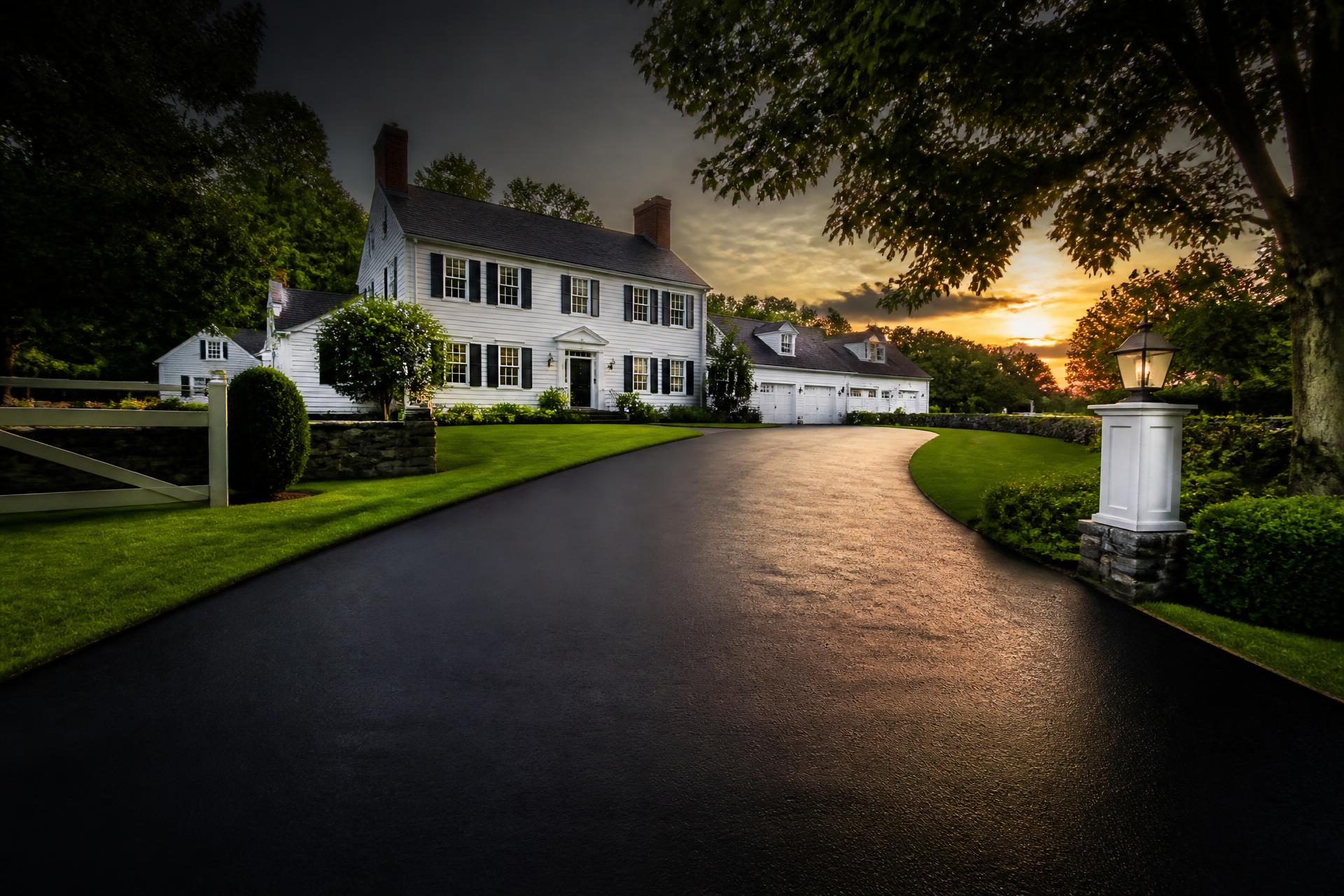 Freshly sealcoated black asphalt driveway leading to a white New England colonial home at sunset
